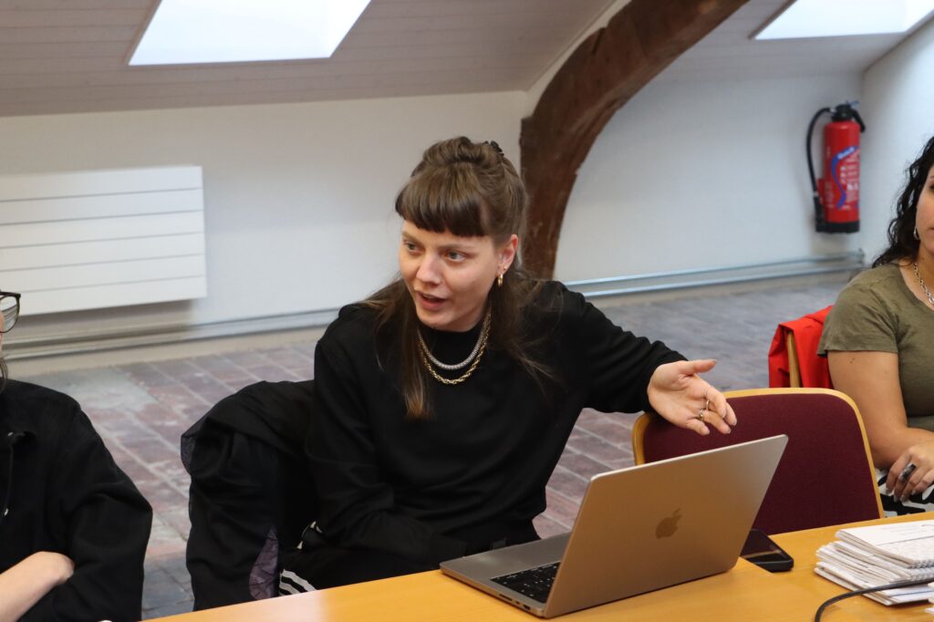 Une femme est assise à une table où se trouve son ordinateur portable. Elle s'exprime, avec la main gauche ouverte, posée sur le dossier de la chaise à sa gauche. Elle porte un pull noir et un collier.